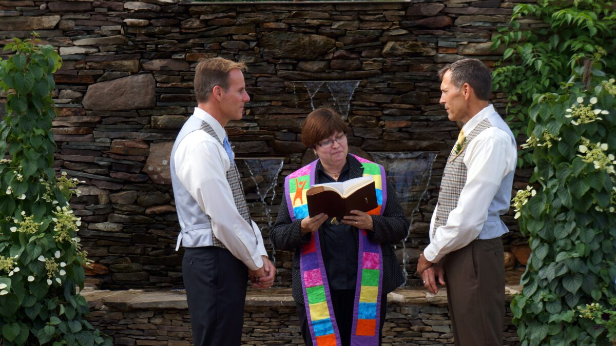 Humanist Celebrant Charlene Komar facilitating a ceremony between two men, in front of a rocky waterfall structure.